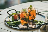 Summer refreshing cold peach ice tea with fresh mint in glass jars on metal tray over wooden garden table