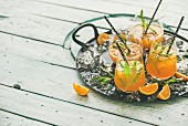 Refreshing cold alcoholic summer citrus cocktail with orange, peppermint and crushed ice in stemless glasses on dark tray