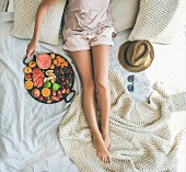 Junge Frau in Shorts relaxed mit Tablett voller frischer Früchte auf Bett (Aufsicht)