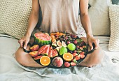 Junge Frau in Shorts sitzt mit Tablett voller frischer Früchte auf Bett