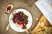 Beetroot and mint salad on a white plate with vintage cutlery and torn crusty bread on an olive wood chopping board, book, salt and pepper pots