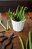 Green beans stacked in a white tin, with scissors resting beside on a rustic tabletop