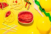 Watermelon cut in half with yellow plates, tablecloth, cutlery and balloons, along with streamers and straws