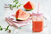 Watermelon smoothie in a jug with a straw in front of a plate of melon slices and fresh mint