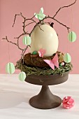 Twigs with paper decorations around ostrich egg on cake stand