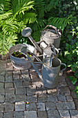 Old zinc watering cans and planted sieve on cobbles