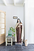 Large statue of the Madonna next to demijohn used as vase on stool