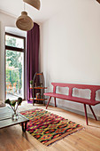 Red-painted wooden bench, vintage shelves, low coffee table and colourful rug in front of terrace doors in living room