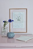 Gesticktes Blumenmotiv im Rahmen hinter Glas, davor Kornblumen