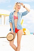 A young blonde woman on a beach wearing a bikini and a denim shirt