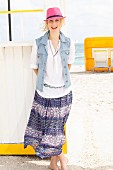 A young blonde woman on a beach wearing a pink hat, a white blouse, a denim gilet and a purple summer skirt