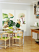 Wooden chairs with turned legs and green seat cushions around dining table in front of large botanical artworks