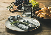 Tischgedeck mit Serviette, Besteck und Olivenzweigen dahinter Windlicht, Semmeln und Trauben