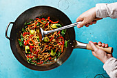 Udon stir fry noodles with oyster mushrooms and vegetables in wok pan and male hands on blue background