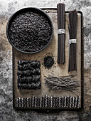 A geometric layout of black pastas (rice, spaghetti, ramen, etc…) on a dark metal surface