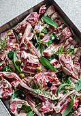 Pieces of raw lamb with garlic, olives, sage, rosemary and bay leaves on a baking tray