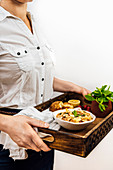 A woman with a white shirt holding a tray with a bowl of cabbage soup, diner rolls, fresh mint leaves