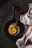 Homemade pancakes with fried orange, served in cast-iron pan on kitchen towel over dark old rusty background
