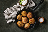 Accordion hasselback baked potatoes served in cast-iron pan with sea salt, cream-fresh and green spring onion on textile napkin