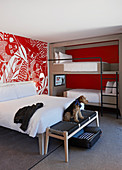 Modernes Hotelzimmer mit Doppelbett, Etagenbett und Hund auf Kleiderbank, rot-weiße Tapete an der Wand