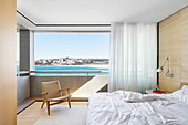 Doppelbett, Wandleuchte und Designer Armlehnstuhl vor Panoramafenster mit Meerblick