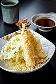 Ebi and yasay tempura with vegetables and prawns deep fried in a light tempura batter on a plate and black table served with a dashi based dipping sauce in the background and green tea