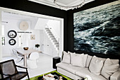 Light upholstered sofa with pillows, large-format picture with sea motif in the living room with black walls, white dining room in the background
