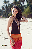 A young brunette woman on a beach wearing a brown bathing suit and harem trousers