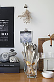 Kitchen utensils, cutlery in glasses, books and pebbles on wooden worksurface