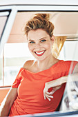 A blonde woman sitting in a car wearing an orange blouse