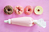 Mini-Donuts mit Schokolade, Zuckerglasur und Zuckerstreuseln in einer Reihe, davor Spritzbeutel