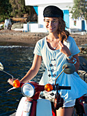 A young woman on a motor scooter wearing a white top, shorts and a black hat