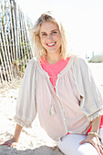 Junge blonde Frau in weißer Bluse und rosa Unterziehshirt am Strand sitzend
