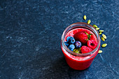 Jar of berry detox smoothie with frozen berries and pumpkin seeds over dark concrete background