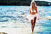 A young blonde woman on a beach wearing sunglasses and a white bikini