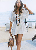 A young woman on a beach wearing a short, white summer dress and a necklace