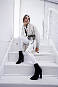 A young woman sitting on a flight of steps wearing a white cocoon coat, white trousers and black boots