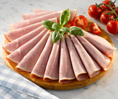 Slices of honey roasted ham on a circular wooden cutting board with garnish of basil and tomatoes