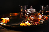Small glass teapot and glasses with hot black tea, dried rose petals, pocket magnifier on golden chain, squeezed orange slice