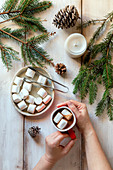 Hands holding a cup of hot chocolate, white candle, spruce cones and spruce tree branches on the table