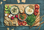 Various Vegetarian dips: hummus, babaganush and muhammara with crackers, bread, fresh vegetables on wooden board