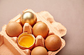 Sechs Hühnereier in Eierkarton, ein goldenes Ei und ein aufgeschlagenes Ei