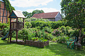 Wooden playhouse with slide, wicker raised beds and seating area below tree in garden