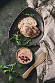 Chicken homemade liver pate in glass jar with sliced whole grain bread, wood knife, cranberries, green salad served on ceramic plate