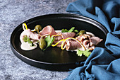Vitello tonnato italian dish. Thin sliced veal with tuna sauce, capers and coriander served on black plate with blue textile