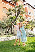 Zwei junge Frauen mit Hut im Spitzenrock und Spitzenkleid machen Selfie im Garten