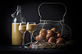 Homemade egg liqueur next to an old wire basket with fresh eggs on straw
