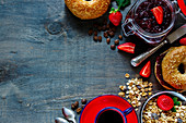 Top view of coffee cup, granola, fresh bagel with jam and strawberry for breakfast on rustic wooden background