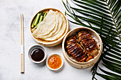 Sliced Peking Duck in bamboo steamer served with fresh cucumber, roasted wheaten chinese pancakes and sauce Hoysin on white wooden background