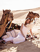 A young woman wearing a long white wedding dress reclining against two camels in the desert